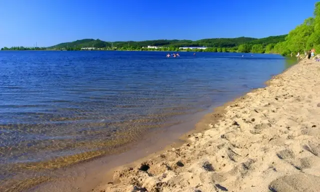 Słoneczny dzień nad zalewem Pilica. Piaszczysta plaża, spokojna woda i zielone wzgórza w tle. Kilka osób pływa i odpoczywa.