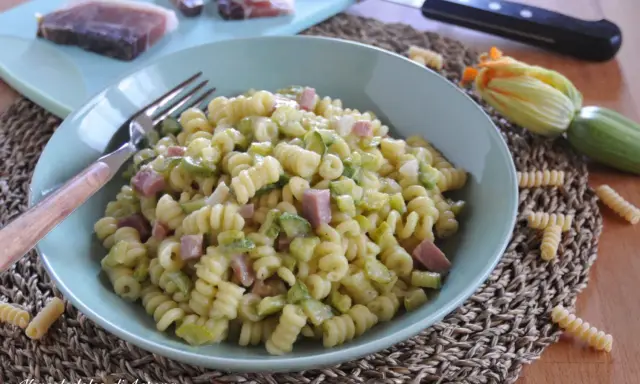 Piatto di pasta fusilli con zucchine e cubetti di speck, guarnito con un fiore di zucca e ingredienti freschi sul tagliere.