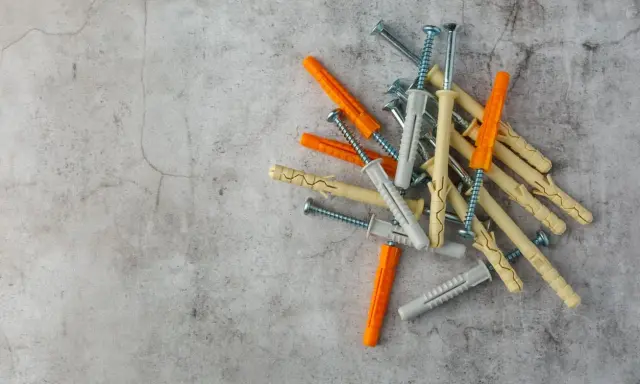 Una tabla de tacos y tornillos de diferentes colores (naranja, gris y beige) sobre una superficie de cemento.