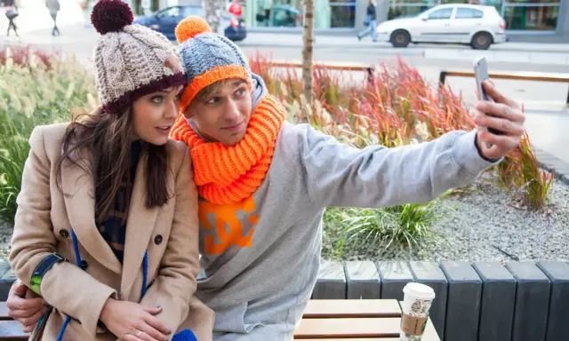 Para do selfie w zimowych stylizacjach. Ona w beżowym płaszczu i czapce z pomponem, on w szarej bluzie i czapce z pomarańczowym akcentem. Jaka czapka do płaszcza? Ta jej wygląda świetnie!