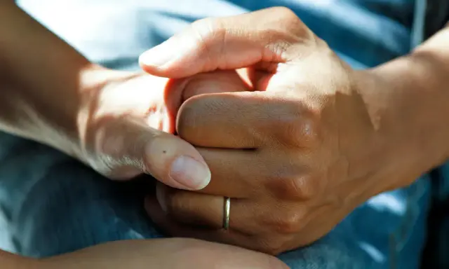 Två händer håller varandra, en gest som visar medlidande och stöd. En tunn ring syns på ena handen.