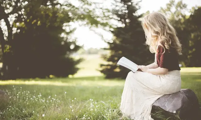Kobieta z blond włosami czyta psalm 6 tekst, siedząc na kamieniu w trawie, otoczona zielenią.