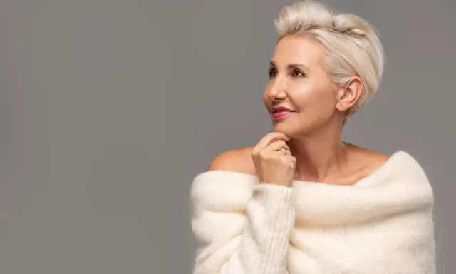 Le visage d'une femme de 80 ans, aux cheveux blonds courts, porte un pull blanc en maille. Elle regarde sur le côté, la main sous le menton.