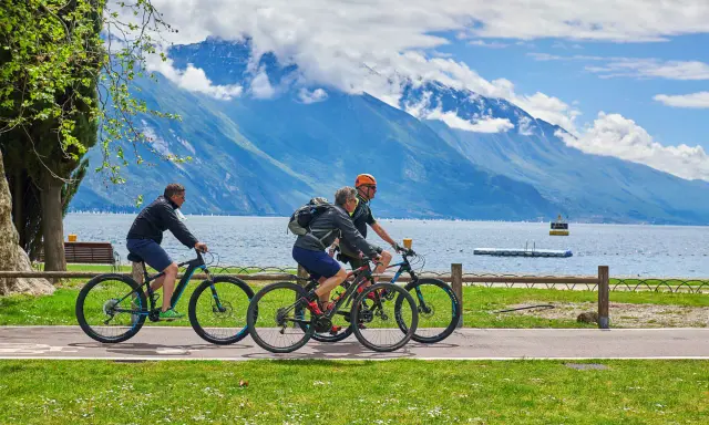 Ciclisti godono di una giornata soleggiata lungo uno dei laghi nord Italia, con montagne maestose sullo sfondo.