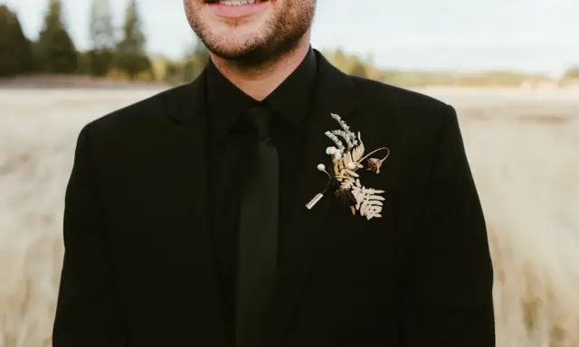 A groom in all black wedding guest attire men's suit, complete with a black tie and a rustic boutonniere.