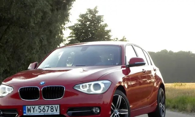 Czerwone BMW 118d na tle drzew i pól.