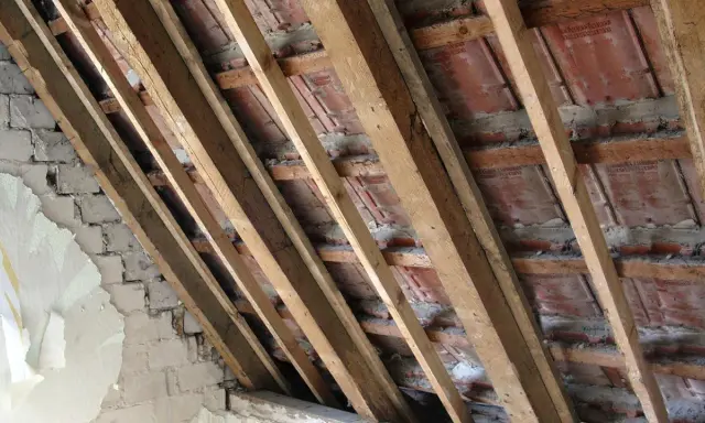 Charpente en bois avec des tuiles rouges et un mur en briques décrépit. Les contre latte sont visibles, soutenant la structure.