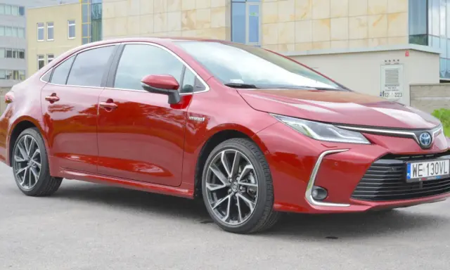 Błyszcząca, czerwona Toyota Corolla 1.8 Hybrid stoi na tle nowoczesnego budynku.