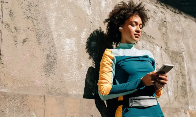 Frau mit Kopfhörern lehnt an Wand, hält handy beim joggen. Musik und Motivation für die Laufstrecke.