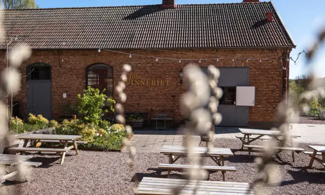 Utomhusmiljö vid Lilla Spinneriet med tegelbyggnad, trädgård och flera picknickbord.