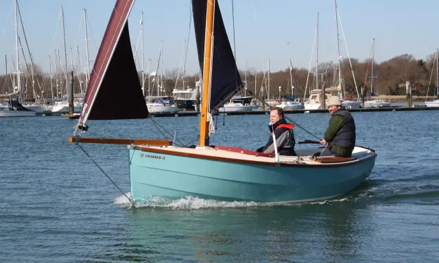 Deux hommes naviguent sur un petit bateau à voile bleu clair, avec d'autres voiliers en arrière-plan.