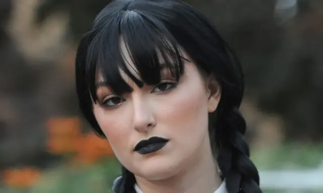 Maquillaje de Miércoles Addams con labios negros y ojos ahumados, peinado con trenzas y flequillo.
