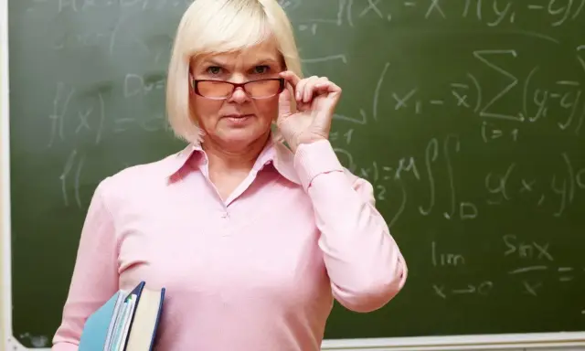 Nauczycielka matematyki poprawia okulary, trzymając książki. Na tablicy widać wzory. Dziś jest 14 października.