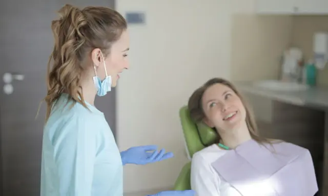 Dentystka w niebieskiej bluzie i rękawiczkach rozmawia z pacjentką. Długie studia stomatologiczne wymagają zaangażowania.