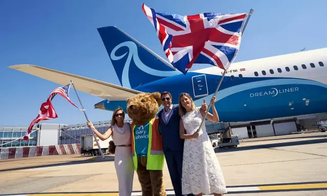 Två kvinnor och en man med en björndräkt firar framför ett flygplan från Norse Atlantic Airways. En kvinna håller en amerikansk flagga, en annan en brittisk flagga.