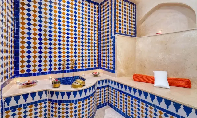 Ein luxuriöser Hamam in Marokko, verziert mit blauen und orangen Zellige-Fliesen. Kerzen und Schalen mit Rosenblättern schaffen eine entspannende Atmosphäre.