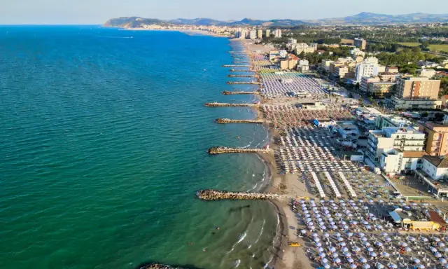 Vista aerea della costa adriatica, con la spiaggia affollata di ombrelloni e stabilimenti balneari, che si estende fino a Cattolica e Misano.