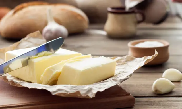 Ein Messer schneidet ein Stück von einem Block welcher Butter. Daneben liegen Knoblauch und ein Brot.