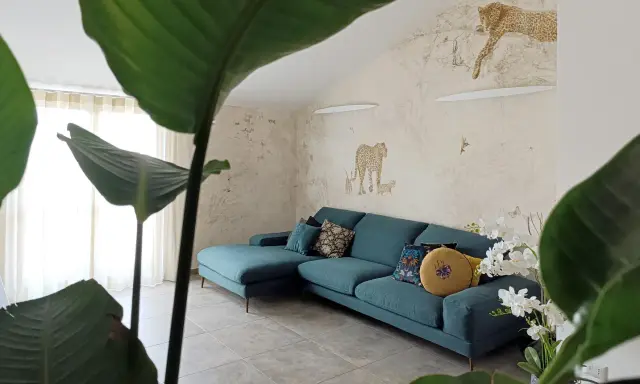 Divano turchese in un salotto con carta da parati a tema giungla e piante rigogliose, per un perfetto **stile jungle chic**.