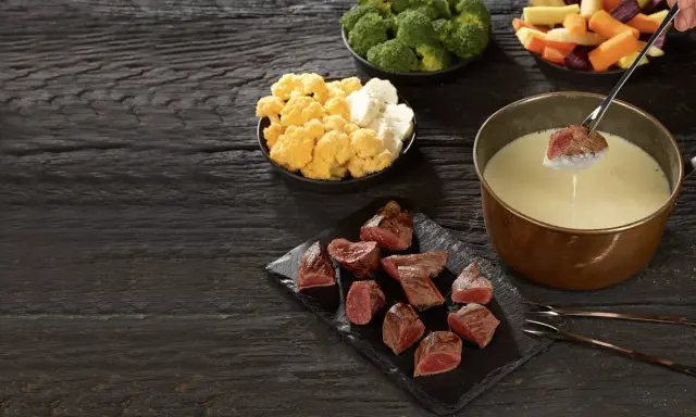 Un morceau de viande est trempé dans un caquelon de fondue. Des morceaux de viande, du brocoli, du chou-fleur et des légumes racines sont disposés sur une table en bois sombre.
