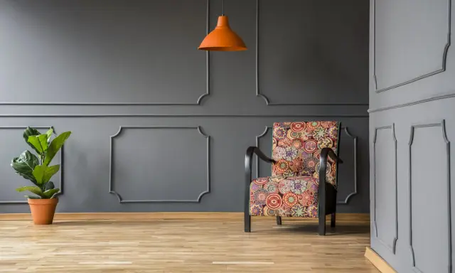 Nowoczesne sztukateria ścienna inspiracje: fotel w kolorowe wzory, pomarańczowa lampa i roślina na tle szarej ściany.
