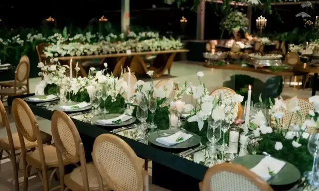 Eleganckie wesele na 100 osób. Długi, lustrzany stół z białymi kwiatami i świecami, otoczony wiklinowymi krzesłami.
