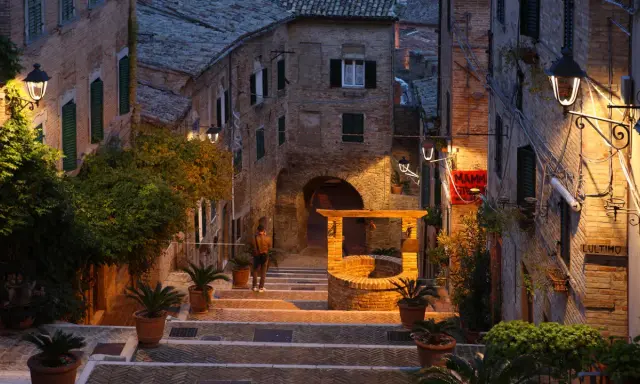 Antica scala a Gabicce Monte illuminata da lampioni, con un pozzo in mattoni e piante in vaso.