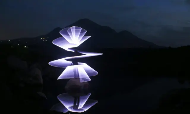 Un artista crea forme luminose con la light painting fotografia, disegnando spirali e petali di luce viola su uno sfondo scuro con montagne e riflessi sull'acqua.