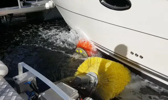 Nettoyage de la partie immergée d'un bateau avec des brosses rotatives jaunes et oranges. L'eau éclabousse autour des brosses.