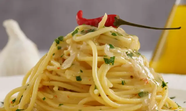 Spaghetti conditi con aglio, olio e peperoncino, guarniti con un peperoncino rosso e prezzemolo. Sullo sfondo, un bulbo d'aglio e una bottiglia d'olio.