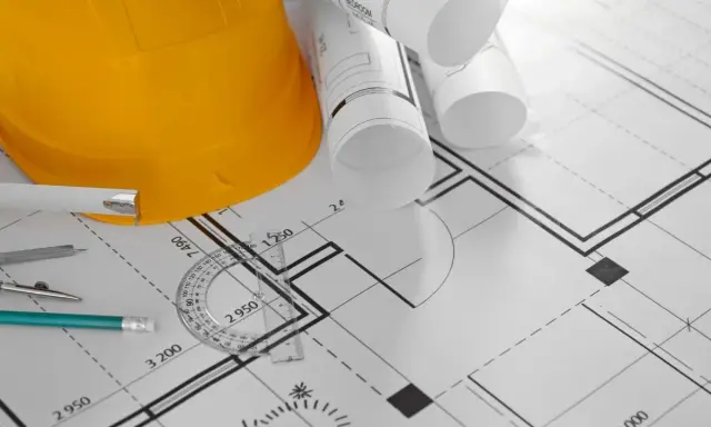 Żółty kask budowlany, zwinięte plany i przybory kreślarskie na projekcie domu. Może posłużyć jako wzór zgłoszenia samowoli budowlanej.