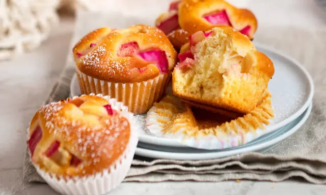 Saftige Rhabarber Muffins, mit Puderzucker bestäubt, liegen auf einem Teller. Einer ist angeschnitten und zeigt die rosa Rhabarberstücke.