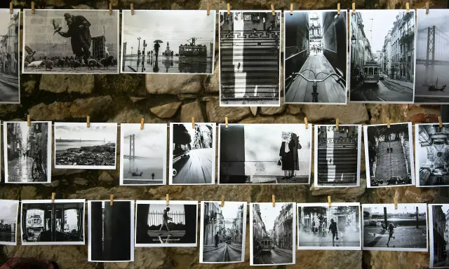 Czarno-białe fotografie wiszą na sznurku na kamiennej ścianie. Widać na nich uliczne sceny z Lizbony, w tym tramwaje, most i ludzi.