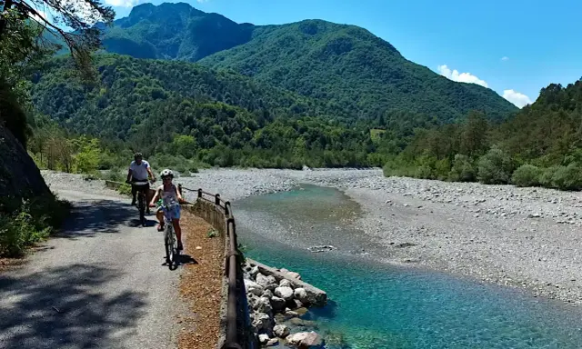 Ciclovia del Tagliamento: due ciclisti esplorano il sentiero panoramico lungo il fiume, circondati da montagne verdi e un cielo azzurro.