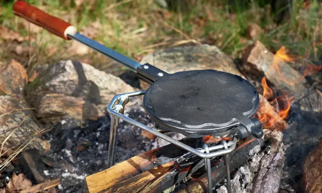 En gjutjärnspanna med trähandtag står på en ställning över en öppen eld. Perfekt för att steka plättar eller våfflor ute i naturen.