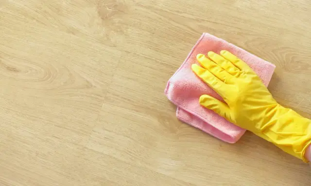En hand i gul handske torkar av ett ljust trägolv med en rosa trasa. Perfekt för pigmenterad olja till ekbord.