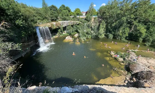 Gente disfrutando de un día soleado en las pozas del río Tus, con una cascada y vegetación exuberante.