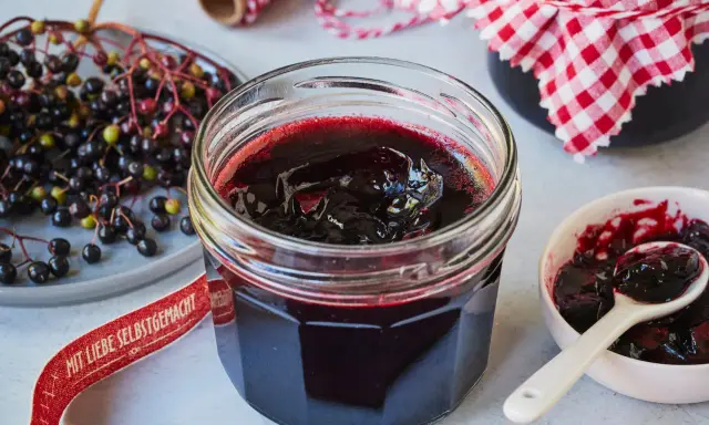 Frisch zubereitete holunderbeeren marmelade im Glas, daneben frische Beeren und eine kleine Schale mit Löffel.