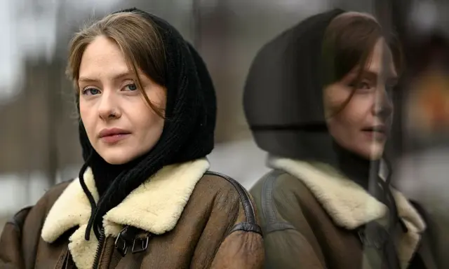 Sofia Kappel i en brun fårskinnsjacka med svart luva, blickar ut. Hennes spegelbild syns i en reflekterande yta.
