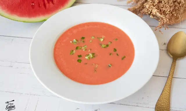 Un refrescante gazpacho de sandía, adornado con trocitos verdes, listo para disfrutar junto a una rodaja de sandía y una cuchara dorada.