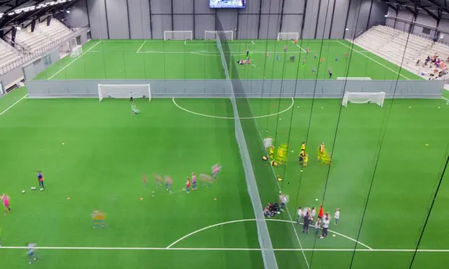 Barn tränar fotboll på en grön konstgräsplan i Prioritet Serneke Arena. Publik sitter på läktaren.