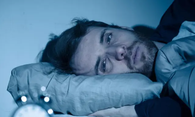 Mann liegt wach im Bett, Augen weit offen, die Angst vor Schlaflosigkeit ist ihm ins Gesicht geschrieben. Ein Wecker ist im Vordergrund zu sehen.