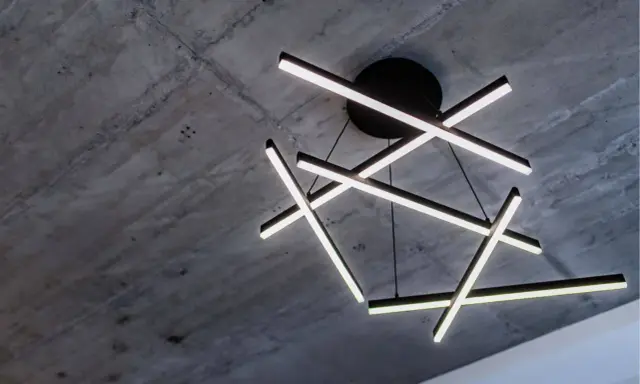Oryginalna lampa sufitowa z geometrycznymi, podświetlanymi ramionami, zawieszona na tle surowego, betonowego stropu.