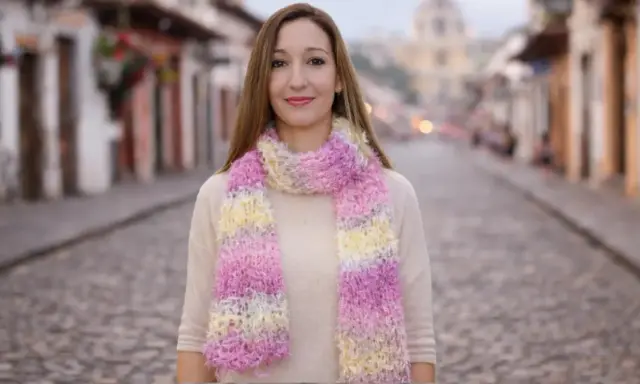 Mujer sonriente luce bufandas de dos agujas modernas en tonos pastel, paseando por una calle empedrada.