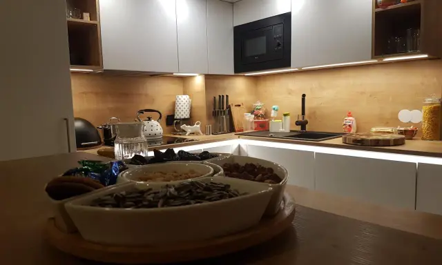 Nowoczesna kuchnia z białymi frontami i drewnianym blatem. Na blacie czajnik, zlew i naczynia z bakaliami. W tle wbudowany czarny okap, który idealnie komponuje się z całością, pokazując jak obudować komin w kuchni.