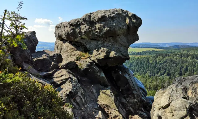 Wielkie, szare skały górują nad zieloną doliną. W oddali widać pola i lasy, a na niebie błękit i chmury.