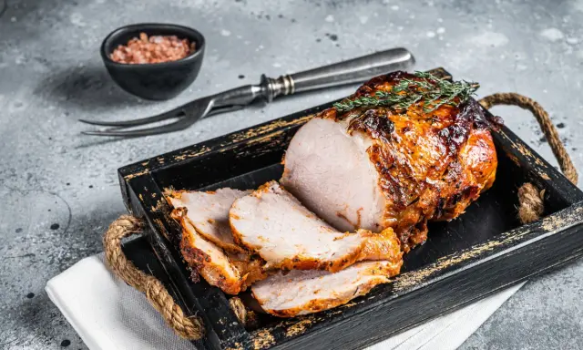 A sliced, roasted pork loin with a golden-brown crust, garnished with thyme, on a rustic wooden tray.