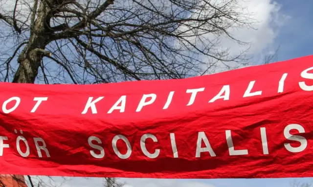 Röd banderoll med vit text: "MOT KAPITALIS FÖR SOCIALIS". En demonstration som visar vad socialism betyder.