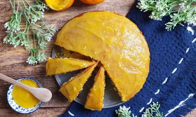 Una torta all'arancia morbidissima, con glassa lucida e profumata, pronta per essere gustata. Accanto, una ciotolina con crema all'arancia e un cucchiaino di legno.