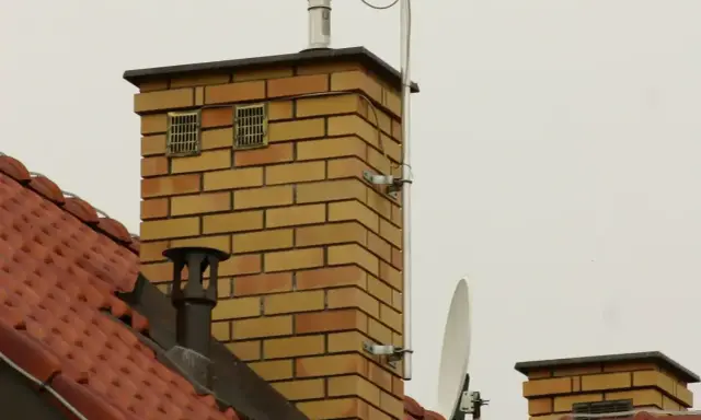 Dwa kratkowane otwory wentylacyjne na ceglanym kominie, zapewniające prawidłowe odpowietrzenie pionu sanitarnego zgodnie z normą.